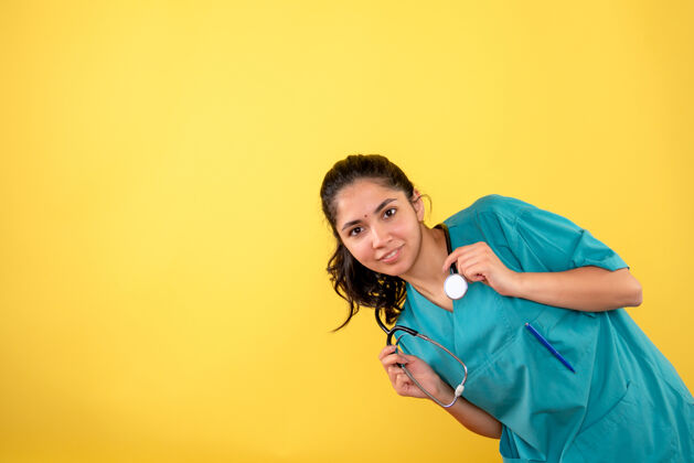 成人黄色墙上带听诊器的年轻女医生的正面图前面快乐微笑