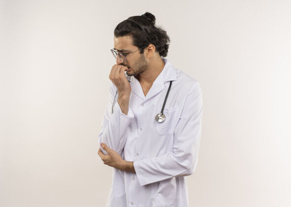 眼镜站在剖面图上的年轻男医生戴着眼镜 穿着白色长袍 手持听诊器 在隔离的白色墙壁上咬着钉子 留着复印空间咬听诊器年轻