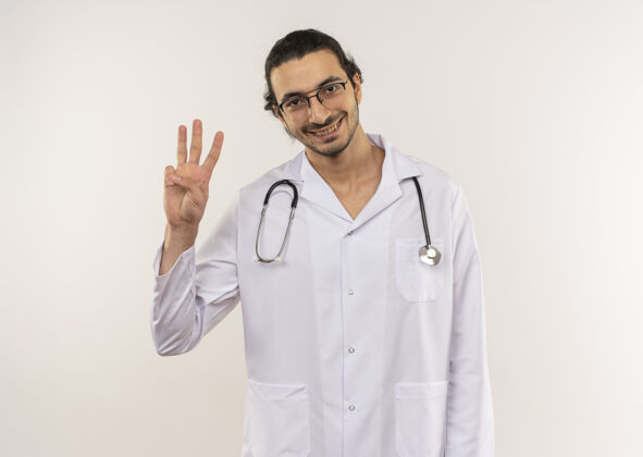 年轻微笑的年轻男性医生 戴着眼镜 穿着白色长袍 听诊器 在隔离的白墙上显示三个 还有复印空间穿长袍男性