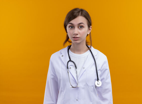 女人穿着医用长袍和听诊器的年轻女医生惊讶地站在隔离的橙色墙上 还有复印空间长袍穿年轻人