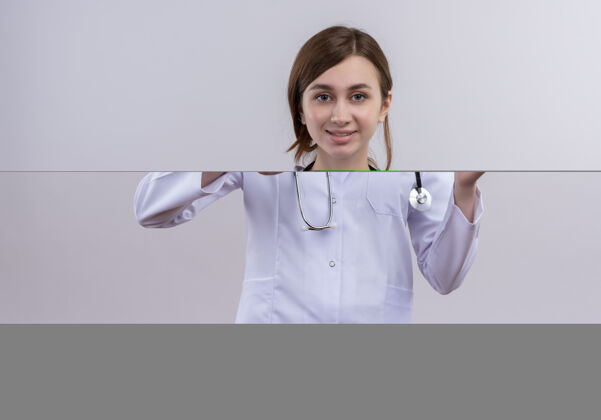 拇指面带微笑的年轻女医生 穿着医用长袍 手持听诊器 竖起大拇指 在隔离的白色墙上拿着带问号的纸条展示听诊器穿