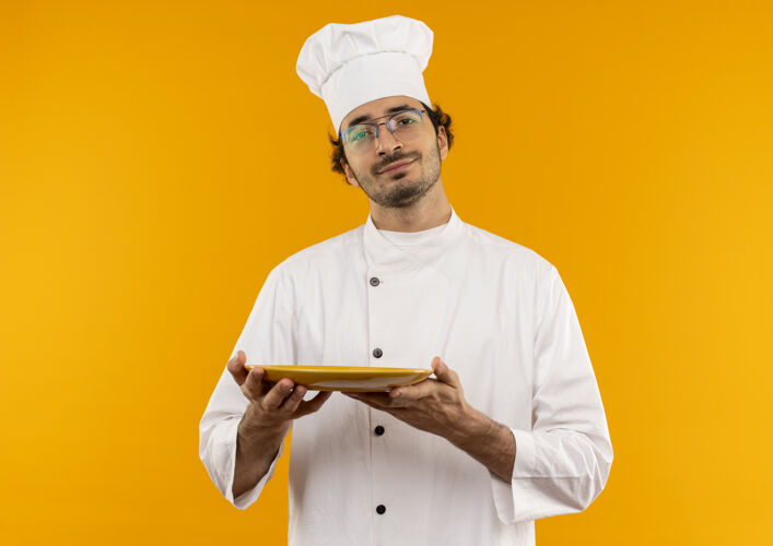 男年轻的男厨师穿着厨师制服 戴着眼镜 把盘子隔离在黄色的墙上年轻制服烹饪