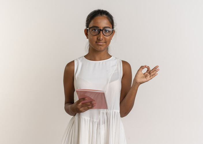 好的高兴的年轻女学生戴着眼镜拿着笔记本 在白色的墙上孤立地展示着奥基的手势拿着眼镜展示