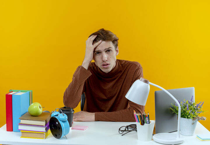悲伤悲伤的年轻学生男孩坐在课桌旁 手放在头上 被隔离在黄色的墙上学生工具头
