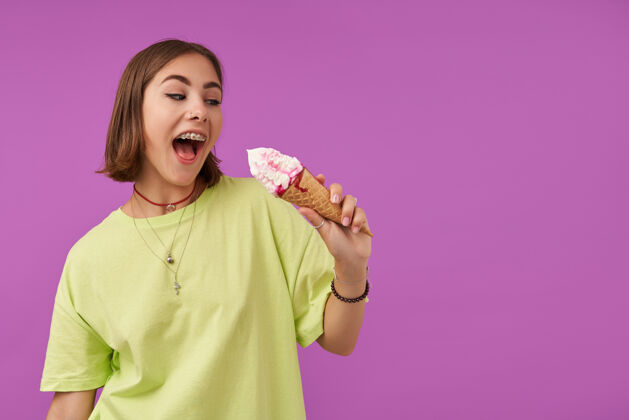 女士照片中 一个漂亮漂亮的女孩手里拿着一个冰激凌 看着右边紫色墙上的复制空间穿着绿色t恤 手镯 戒指和项链时髦成人牙套