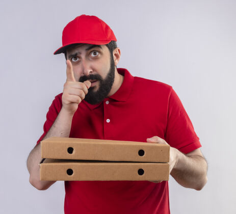 拿着印象深刻的年轻英俊的白人送货员穿着红色制服 戴着帽子 拿着比萨饼盒 指着和仰望孤立的白色帽子盒子红色