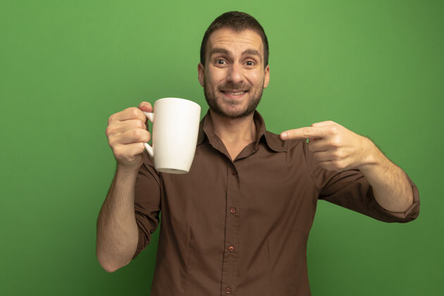 指着年轻的高加索男子微笑着 拿着茶杯 指着绿色背景上的相机杯子茶拿着