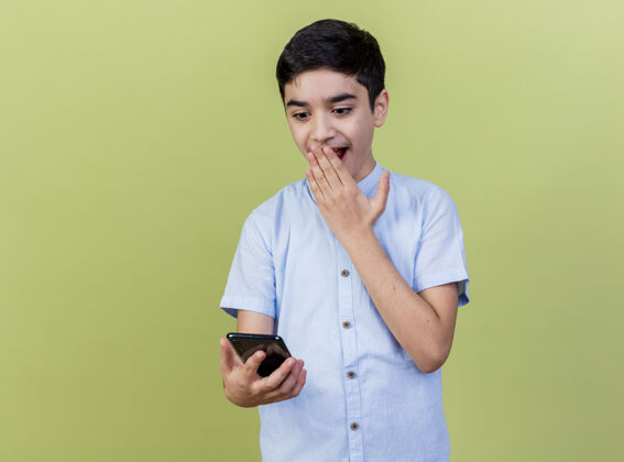电话惊讶的年轻白人男孩拿着手机 看着手机 手放在嘴上 在橄榄绿的背景上 留着空白手机年轻男孩