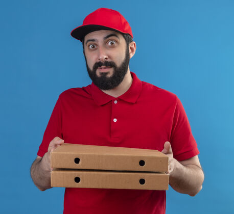年轻印象深刻的年轻英俊的白人送货员穿着红色制服 戴着帽子拿着比萨饼盒孤立在蓝色红色拿着帅气