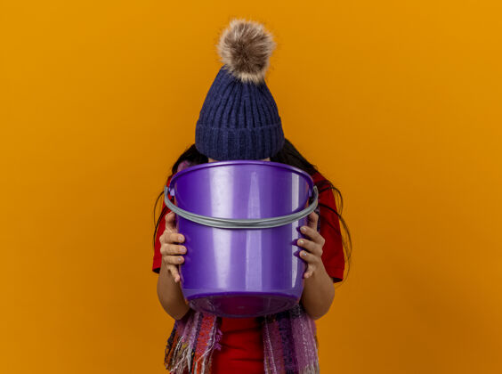 疾病年轻的白种人生病的女孩戴着冬天的帽子和围巾呕吐到塑料桶孤立的橙色背景与复制空间女孩水桶呕吐物