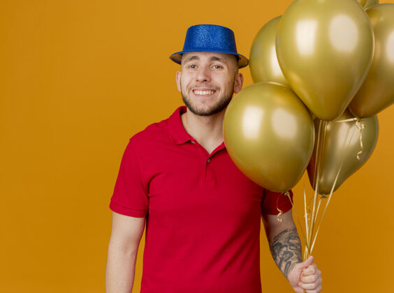 帅气面带微笑的年轻英俊的斯拉夫党人戴着党的帽子拿着气球看着相机孤立的橙色背景与复制空间微笑小伙子抱着