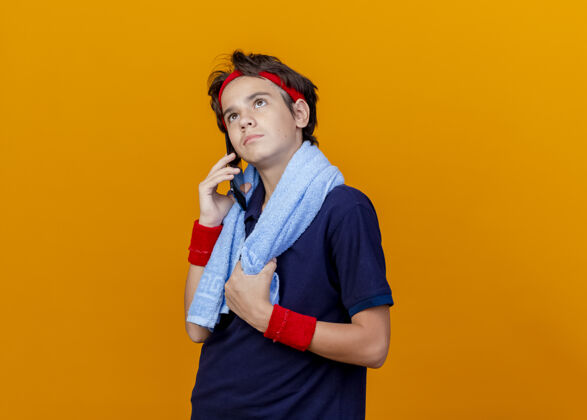 抓住年轻帅气的运动男孩戴着头带和护腕 戴着牙套 脖子上围着毛巾 拿着毛巾在讲电话 在橙色的背景上看着孤立的复制空间电话腕带牙科