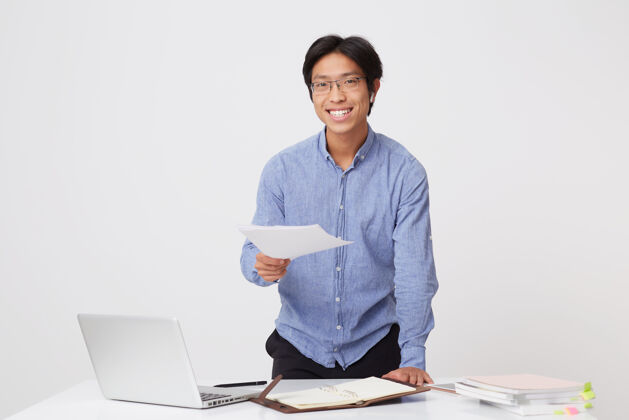 律师带着微笑的亚洲年轻商人 戴着眼镜 穿着蓝色衬衫 戴着耳机 站在白墙上 拿着笔记本电脑和文件工作计划自信文件