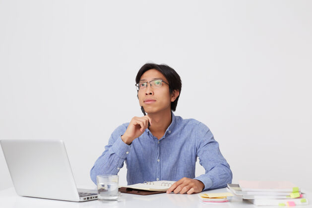 电脑体贴专注的亚洲年轻商人戴着眼镜 在隔着白墙的桌子旁的笔记本上思考和写作办公桌桌子梦想