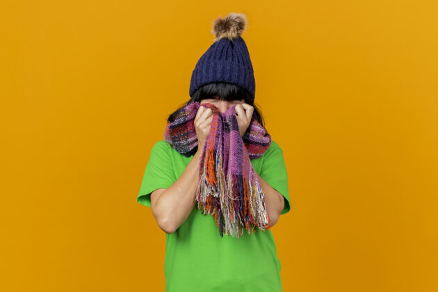 围巾年轻生病的白人女孩戴着冬天的帽子和围巾看着相机拿着围巾在脸上孤立的橙色背景与复制空间帽子年轻生病