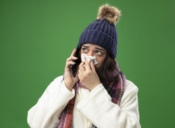 年轻人虚弱的年轻白种人生病的女孩穿着长袍 戴着冬天的帽子和围巾打电话 用餐巾擦鼻子 看着绿色背景上孤立的一面疾病湿巾帽子