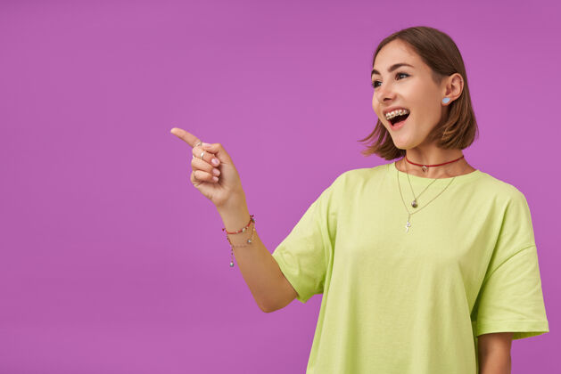 快乐女学生 年轻的女士 笑着用手指指着紫色墙上的复印处展示着一个标志穿着绿色t恤 戴着牙套 手镯和戒指时髦项链女士