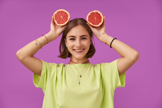 牙齿十几岁的女孩 快乐快乐的微笑着的女士 深色短发 头上戴着葡萄柚站在紫色的墙上穿着绿色t恤 戴着牙套 手镯和项链微笑服装成人