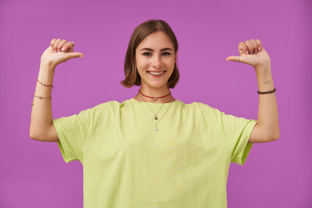 深褐色十几岁的女孩 快乐快乐 深色的短发指着自己的拇指微笑站在紫色的墙上穿着绿色t恤 牙套 手镯和戒指牙套服装牙齿