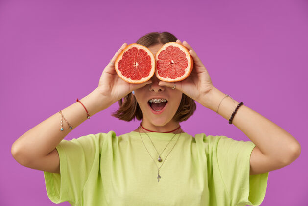 深色十几岁的女孩 快乐快乐 深色短发的女人 眼睛上戴着葡萄柚站在紫色的墙上穿着绿色t恤 戴着牙套 手镯和项链葡萄柚牙齿热带