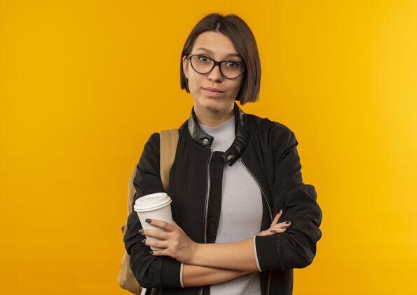 年轻自信的年轻女学生戴着眼镜 背着书包 以封闭的姿势站着 拿着咖啡杯 孤立地放在橙色的空间里橙色立场空间
