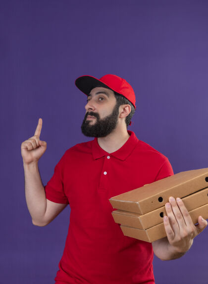 自信自信的年轻英俊的白人送货员身穿红色制服 头戴鸭舌帽 手里拿着披萨盒 一边看着一边 一边指着孤立的紫色穿制服紫色