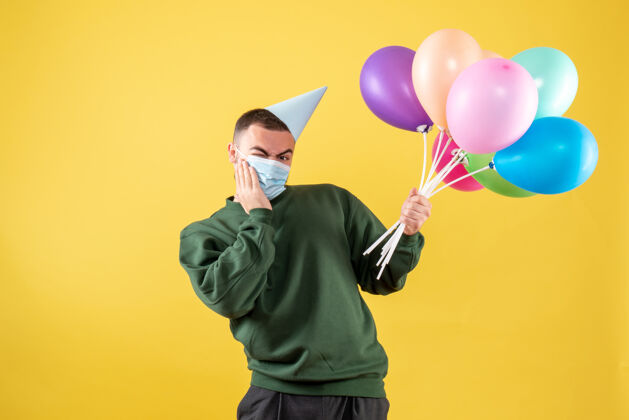 年正面图：年轻男性手持黄色背景上的彩色气球病毒聚会快乐