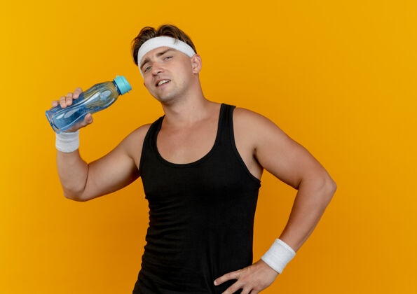 运动自信的年轻帅气的运动型男人戴着头带和腕带 手放在腰上拿着水瓶 孤立在橙色上穿自信腰