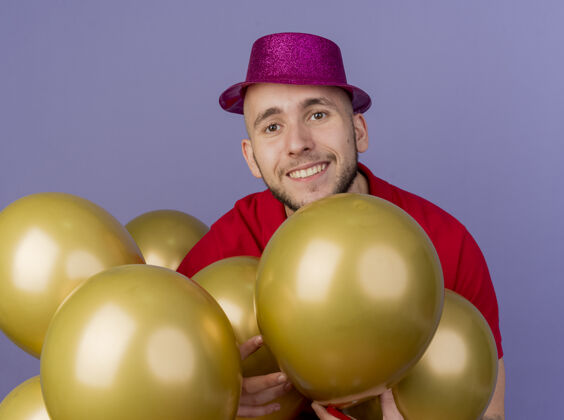 在其中微笑着的年轻英俊的斯拉夫党人戴着党帽站在气球之间触摸他们看着紫色背景上孤立的相机帅气站立帽子