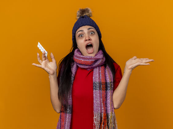 空令人印象深刻的年轻白种人生病的女孩戴着冬天的帽子和围巾 手里拿着一包药片 空手看着隔离在橙色背景上的相机帽子疾病手