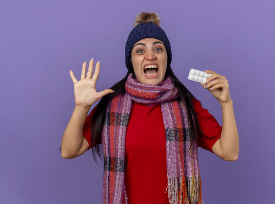 女孩令人印象深刻的年轻白种人生病的女孩戴着冬天的帽子和围巾 展示一包平板电脑 看着相机 显示五只手孤立在紫色背景上药片印象包