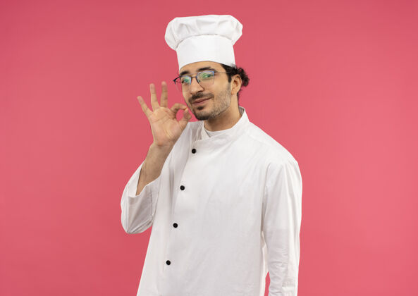 眼镜年轻的男厨师穿着厨师制服 戴着眼镜 在粉红色的墙壁上展示着美味的手势厨师手势表演