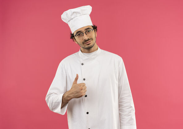 制服年轻的男厨师穿着厨师制服 戴着眼镜 他的大拇指孤立地竖在粉红色的墙上眼镜男厨师