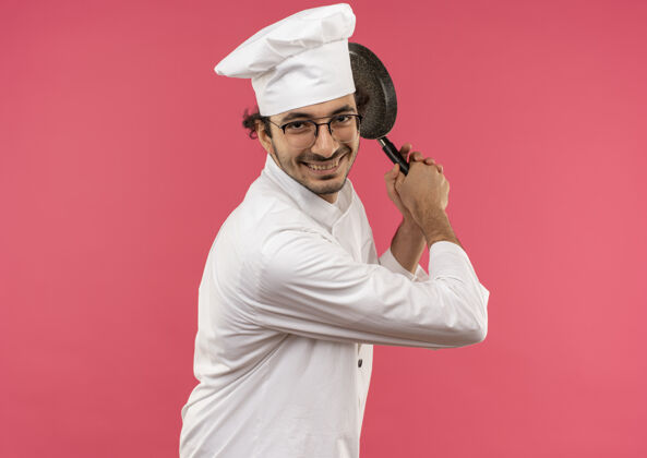 肩膀面带微笑的年轻男厨师穿着厨师制服 戴着眼镜 肩上扛着煎锅 隔离在粉红色的墙上薯条平底锅男