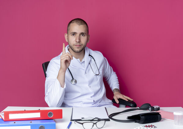 办公室自信的年轻男性医生穿着医用长袍 手持听诊器坐在办公桌旁 手持工作工具 举起手指 把手放在办公椅上 粉色的脸上显得孤立无援自信提高医疗