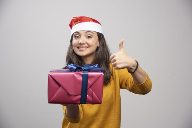 微笑一个戴着圣诞帽的微笑的女人举着一个大拇指 手里拿着一个礼品盒圣诞老人欢呼女士