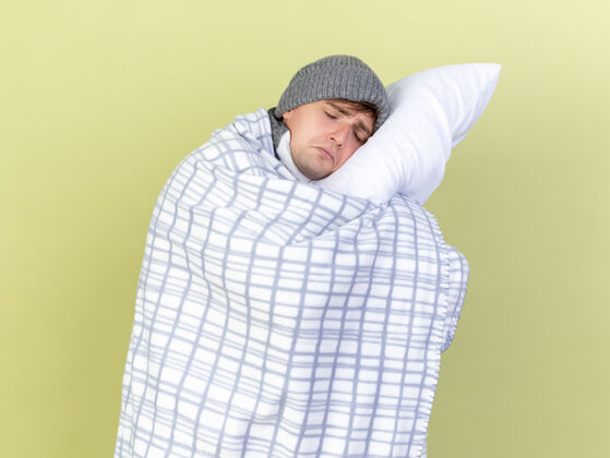 持有悲伤的年轻英俊的金发病男子戴着冬天的帽子和围巾包裹在格子布抱枕上把头放在上面闭着眼睛隔离在橄榄绿的背景上绿色疾病封闭