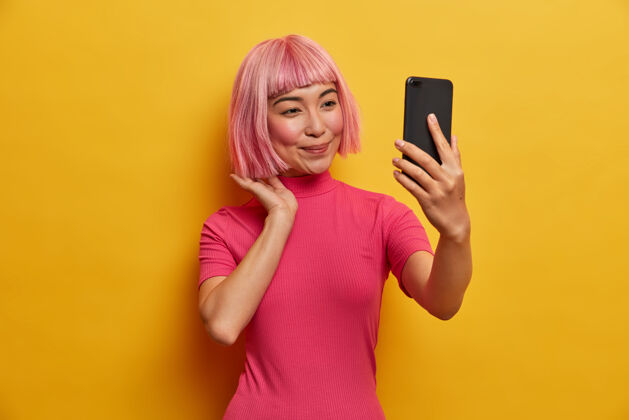 高兴美丽的亚洲女人用视频通话的小玩意 修粉色头发 看智能手机摄像头 自拍粉色发型美丽