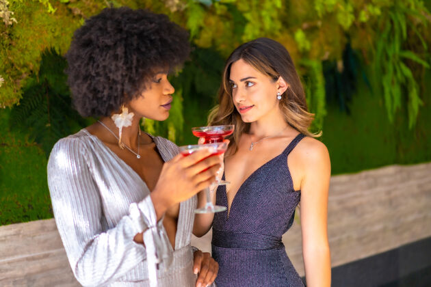 有趣非裔美国人和白人女性朋友穿着花哨的衣服喝酒聊天鸡尾酒人饮酒