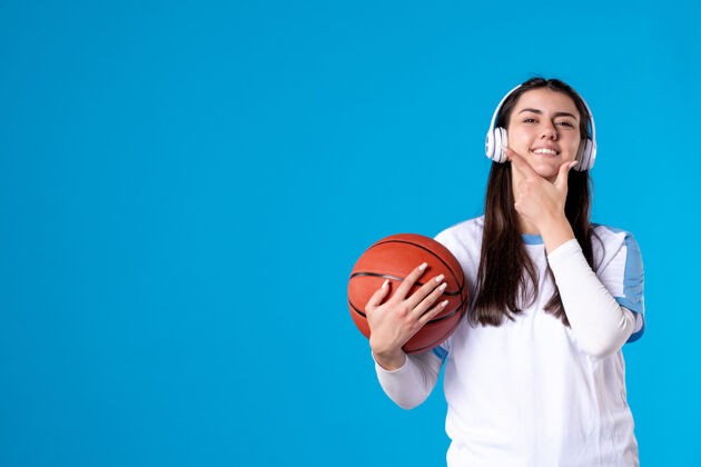 成人正面图：戴着耳机拿着篮球的年轻女性站在蓝色的墙上年轻女性人物年轻人