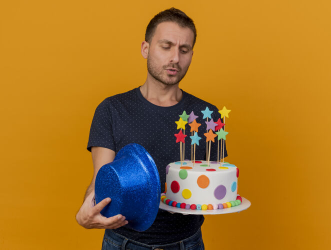 姿势笨手笨脚的帅哥拿着蓝色派对帽 看着橙色墙上孤立的生日蛋糕衣服人不知所措