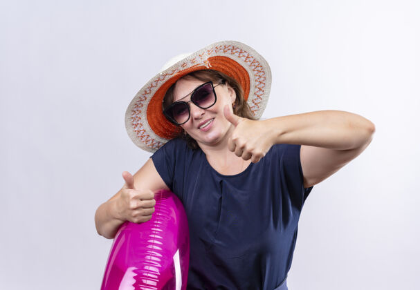 帽子高兴的中年旅行家妇女戴着眼镜 戴着帽子 拿着充气戒指 在孤零零的白墙上竖起大拇指拇指请戴戒指