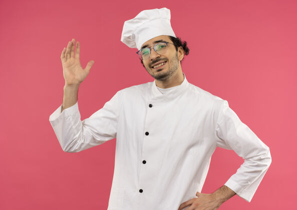 提高年轻的男厨师面带微笑 身穿厨师制服 戴着眼镜 举起手 另一只手放在粉红色的臀部男性厨师另一个