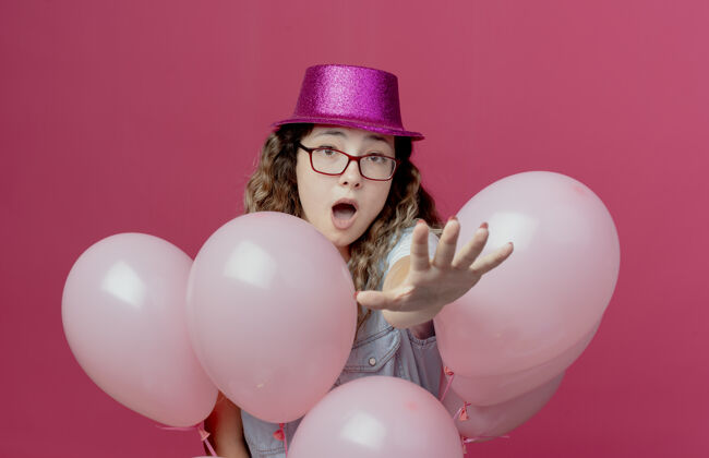 相机那个戴着眼镜和粉色帽子站在气球后面的年轻女孩吓坏了害怕拿着帽子