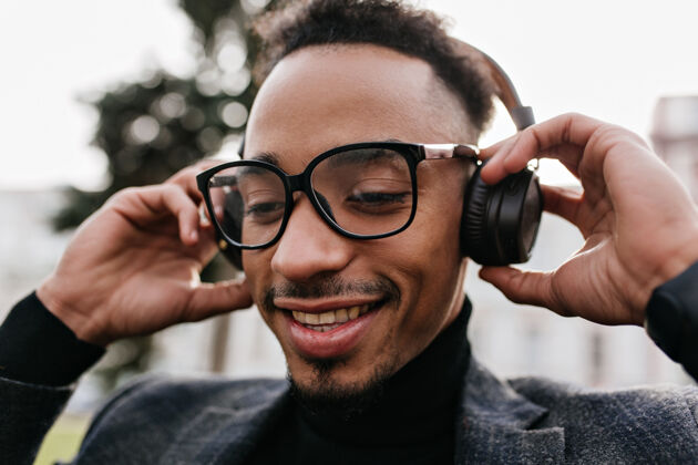 享受公园里戴着休闲眼镜的可爱男模的特写镜头 音乐声震耳欲聋黑头发的非洲帅哥戴着耳机摆姿势的照片男孩城市微笑