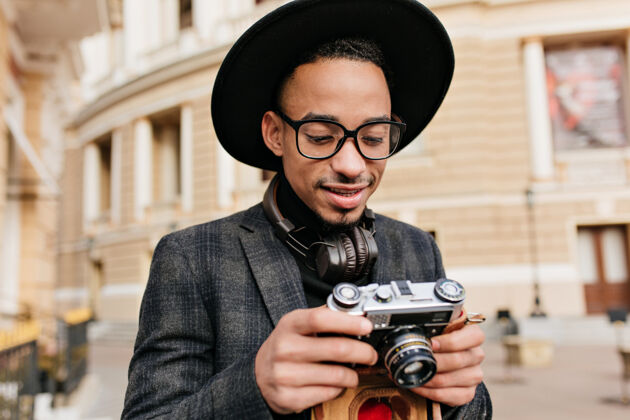 黑发好奇的非洲男人戴着帽子看着相机户外的黑人男摄影师站在美丽的建筑附近的画像夹克表情街道