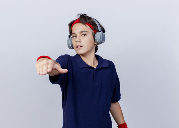 太空自信的年轻帅气的运动男孩戴着头带和腕带 戴着戴着牙套的耳机 看着并指着隔离在白色背景上的相机 还有复制空间白色信心穿