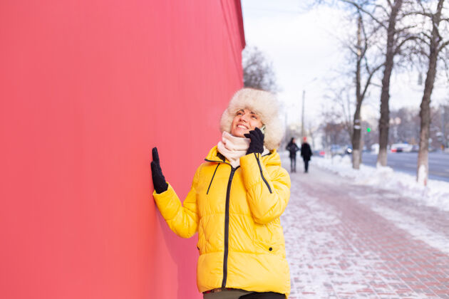 乐趣在一个冬天阳光明媚的日子里 一个穿着暖和衣服的红墙背景上的快乐的年轻女子在雪城的人行道上微笑着打电话通信持有年轻