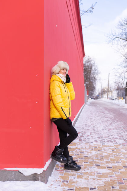 乐趣在一个冬天阳光明媚的日子里 一个穿着暖和衣服的红墙背景上的快乐的年轻女子在雪城的人行道上微笑着打电话持有城市女孩