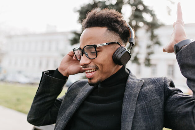 年轻一个留着非洲时髦发型的黑人在城市里听音乐一个穿着休闲夹克戴着耳机的帅哥在公园里摆姿势耳机男孩放松